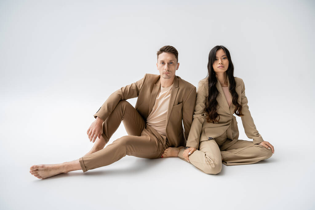 full length of barefoot interracial couple in stylish attire sitting and looking at camera on grey background - Foto, afbeelding