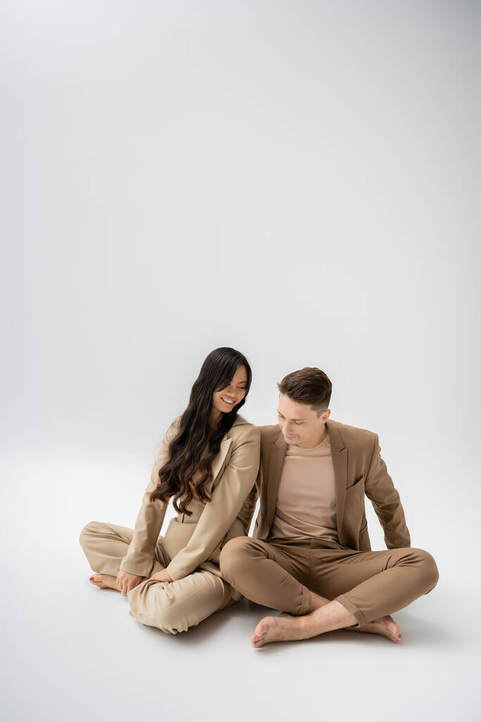 happy interracial couple in stylish pantsuits sitting with crossed legs on grey background - Zdjęcie, obraz