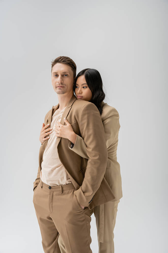 morena asiática mujer abrazando hombre de pie con las manos en bolsillos de pantalones beige aislado en gris - Foto, Imagen