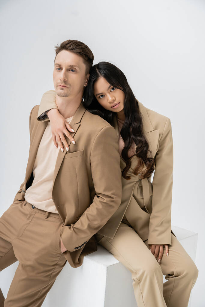 brunette asian woman hugging man in beige suit while sitting on cube and looking at camera on grey background - Valokuva, kuva