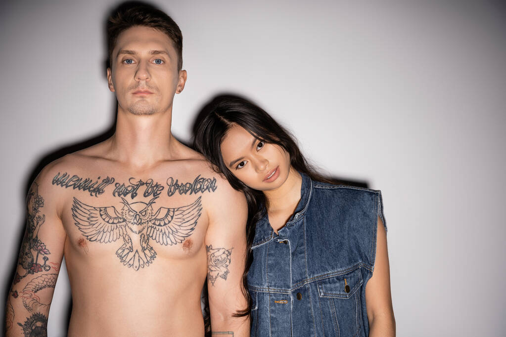 vista frontal de hombre tatuado sin camisa y mujer asiática en chaleco denim mirando a la cámara sobre fondo gris - Foto, Imagen