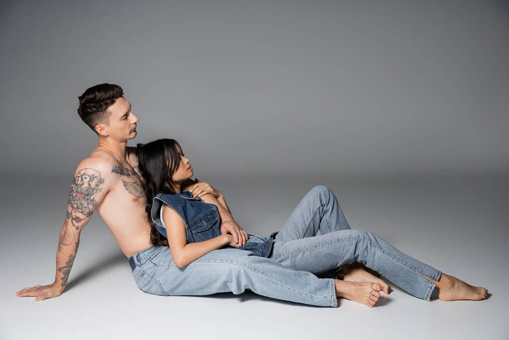 side view of brunette asian woman and shirtless tattooed man in jeans sitting on grey background - Valokuva, kuva