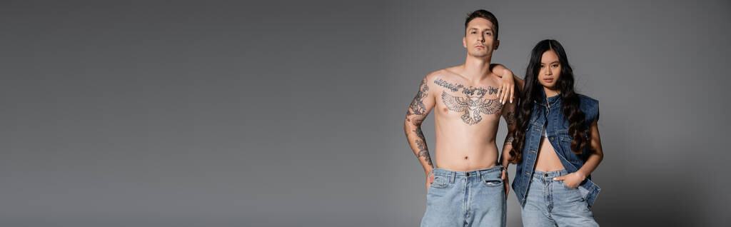 shirtless man with tattooed body and brunette asian woman in denim clothes posing on grey background, banner - Zdjęcie, obraz