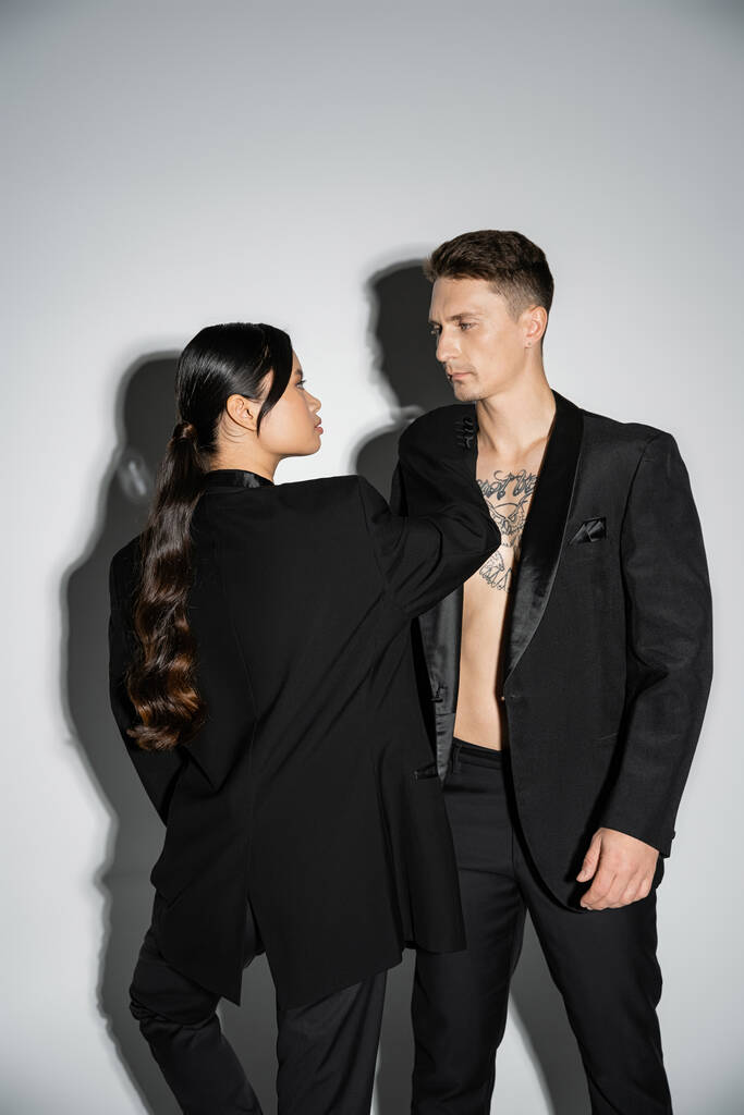 long haired asian woman and tattooed man in black elegant attire looking at each other on grey background - Zdjęcie, obraz