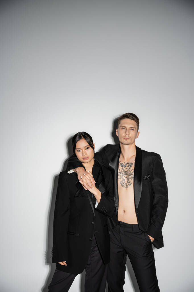 tattooed man in black suit embracing elegant asian woman while posing on grey background - Foto, afbeelding