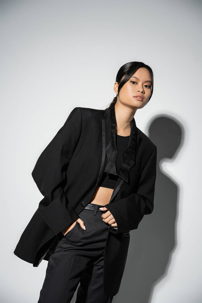 brunette asian woman in black fashionable suit posing with hand in pocket on grey background with shadow - Foto, Imagen