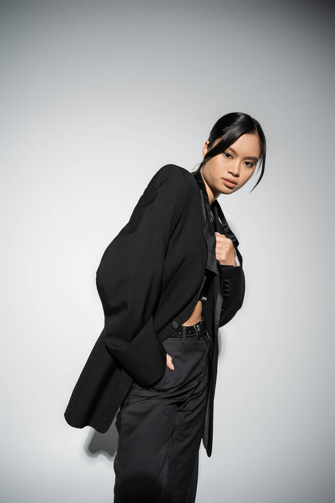 morena mujer asiática en negro elegante blazer y pantalones de pie con la mano en el bolsillo sobre fondo gris - Foto, Imagen