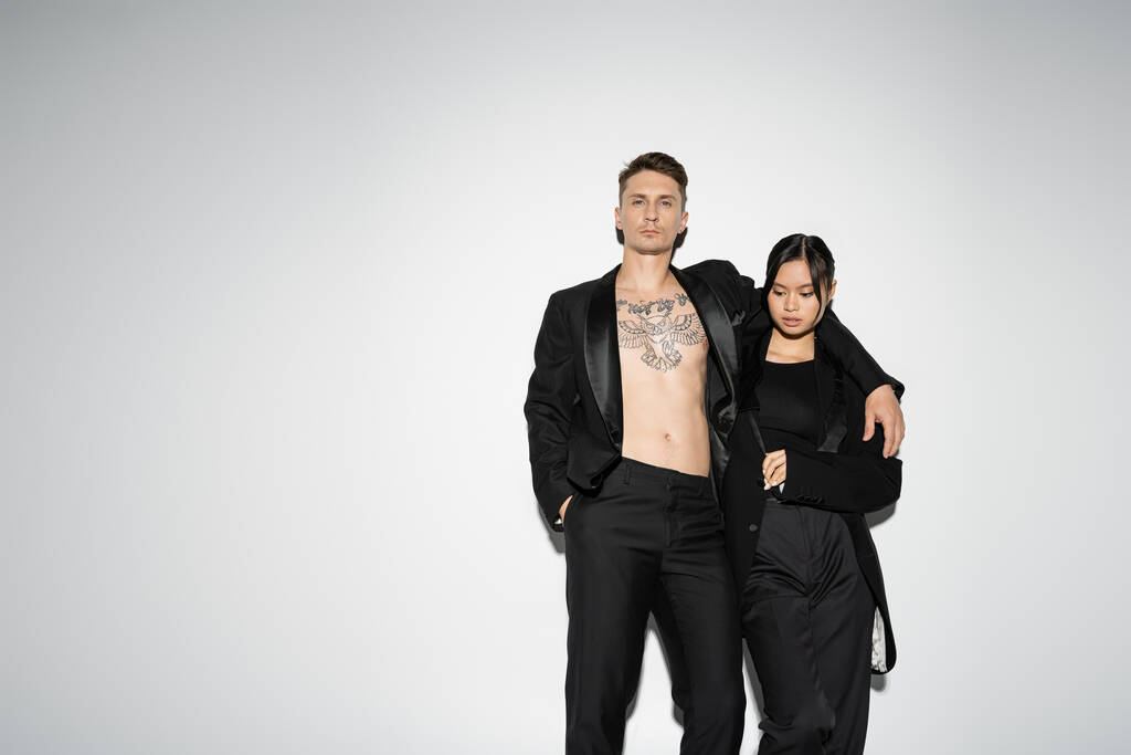 татуйований чоловік в чорному модному костюмі, що обіймає елегантну азіатську жінку, позує з рукою в кишені на сірому фоні
 - Фото, зображення