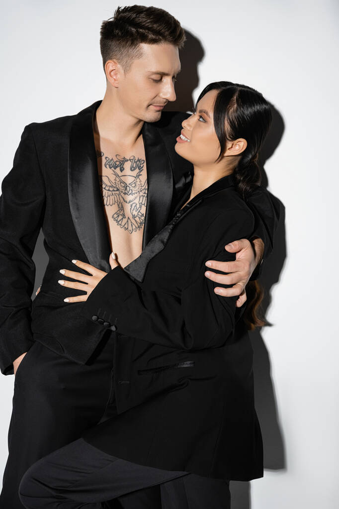 szczęśliwy Azji kobieta i tatuaż mężczyzna w czarny stylowy strój patrząc na siebie na szarym tle - Zdjęcie, obraz