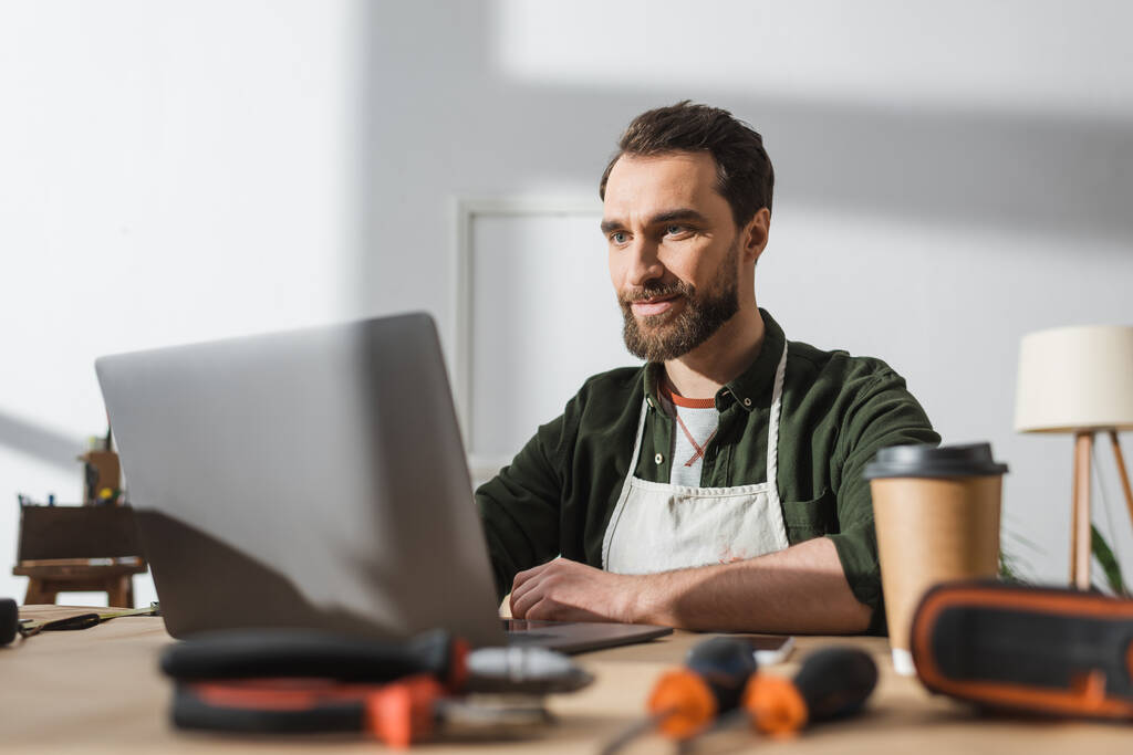 Bearded carpenter using laptop near blurred takeaway coffee and tools in workshop  - Foto, Bild