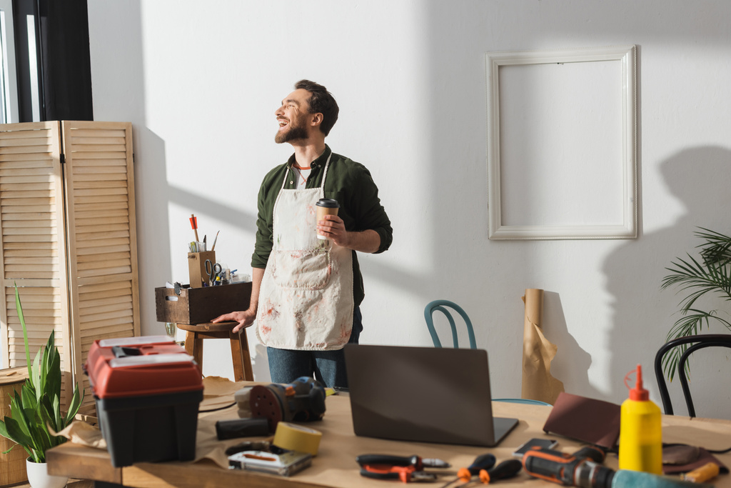 Carpinteiro alegre em avental segurando café para ir perto de ferramentas em oficina  - Foto, Imagem