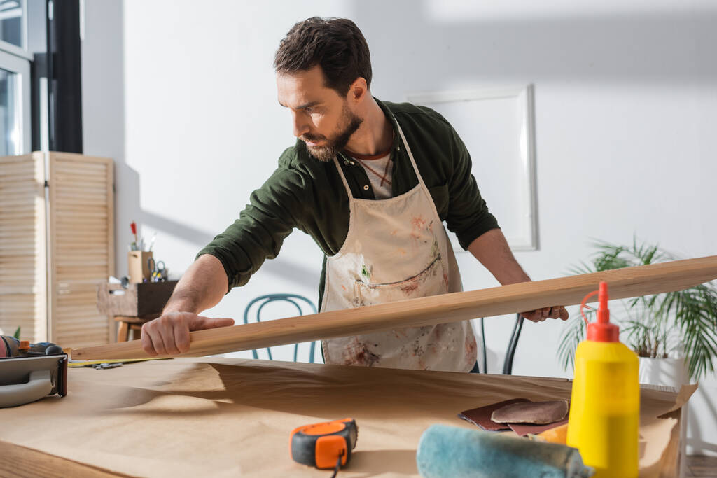 Ξυλουργός σε βρώμικη ποδιά βάζοντας ξύλινη σανίδα στο τραπέζι κοντά χάρακα και γυαλόχαρτο  - Φωτογραφία, εικόνα