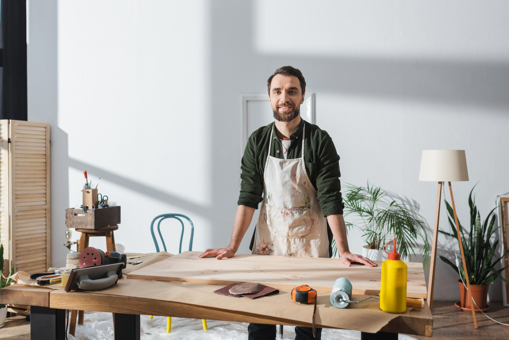 Smiling bearded carpenter in apron looking at camera near wooden board in workshop  - Fotó, kép