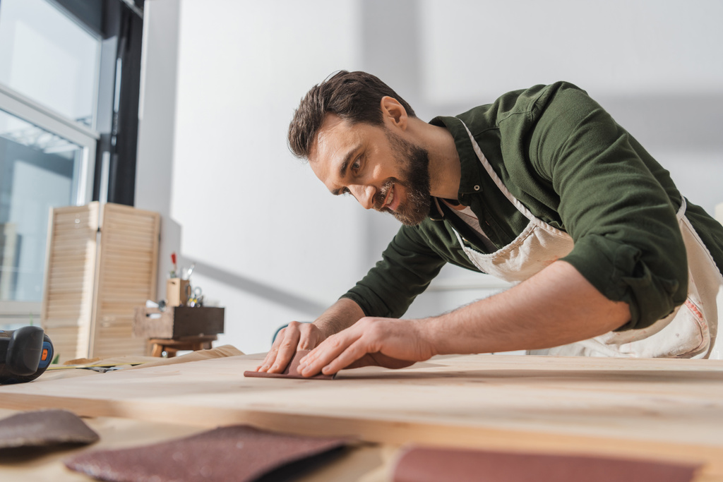 Smiling bearded workman sanding wooden board in workshop  - Fotoğraf, Görsel