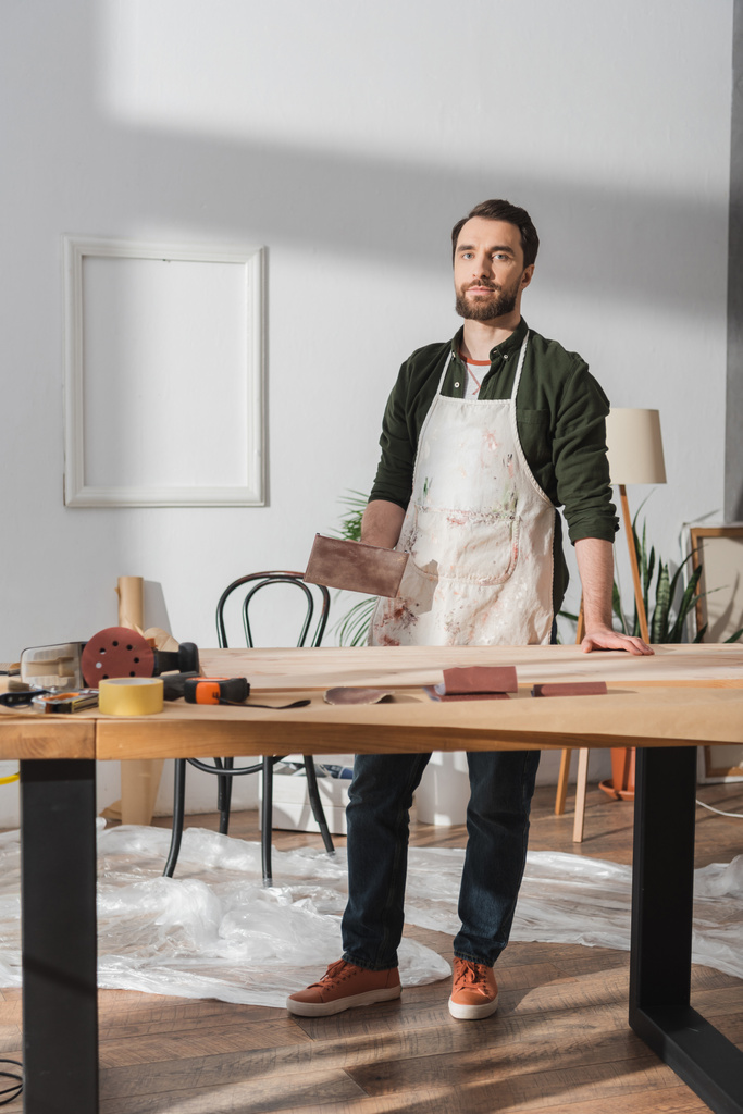 Handwerker in Schürze hält Schleifpapier in der Hand und blickt in die Nähe von Werkzeug und Holzbrett in der Werkstatt  - Foto, Bild