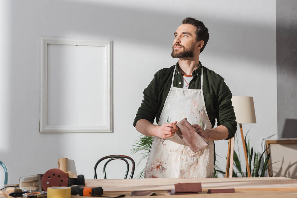 Bearded craftsman in apron holding sandpaper near blurred wooden board on table  - Foto, afbeelding