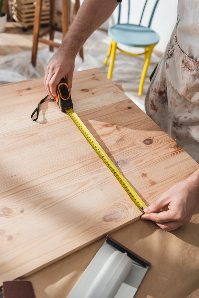 Обрезанный вид плотника в фартуке, измеряющий деревянную доску в мастерской  - Фото, изображение
