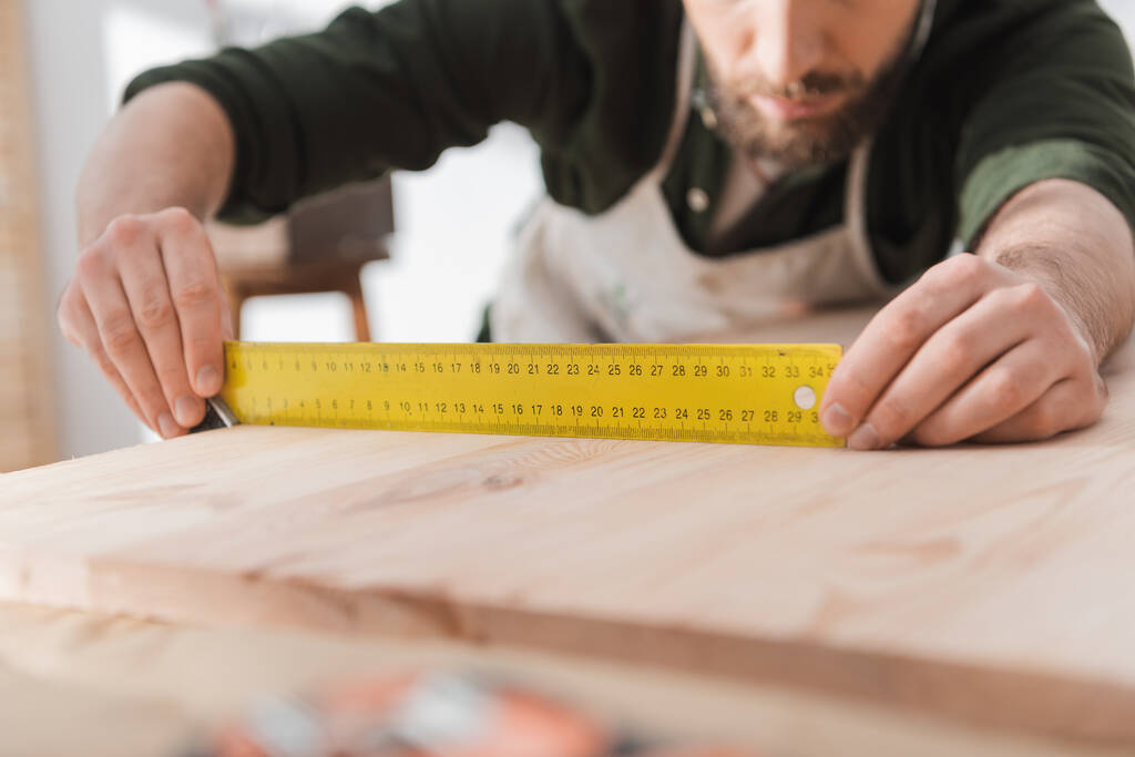 Vista recortada de un artesano borroso sosteniendo una herramienta cuadrada cerca de un tablero de madera  - Foto, Imagen