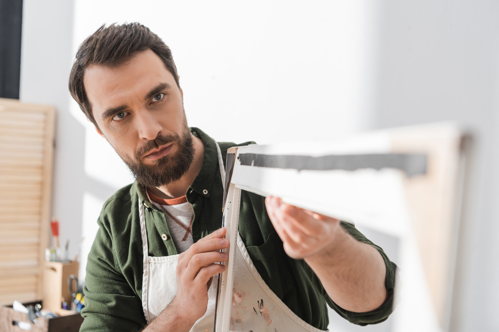 Бородатый ремонтник в фартуке смотрит на размытую деревянную рамку в мастерской  - Фото, изображение