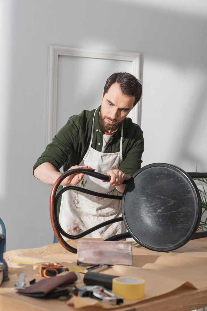 Bearded restorer sanding chair near blurred tools on table in workshop  - Fotografie, Obrázek