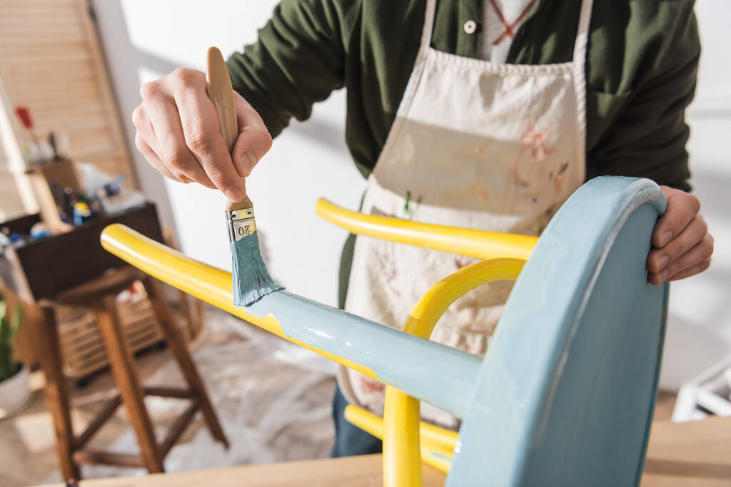 Cropped view of craftsman painting on wooden chair in blurred workshop  - 写真・画像