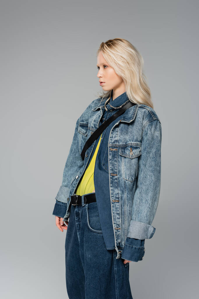 young blonde model in stylish denim outfit looking away while posing isolated on grey  - Photo, image
