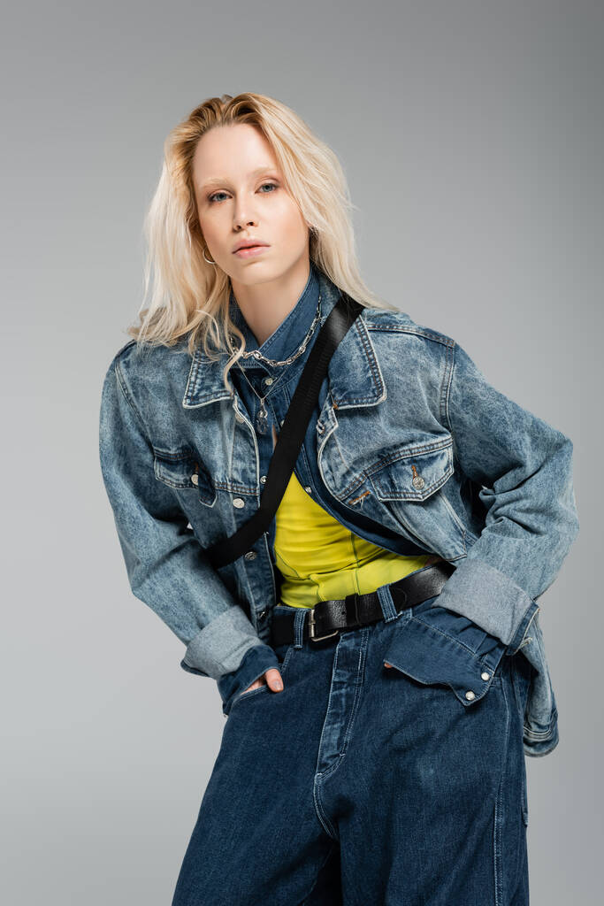young blonde woman in stylish denim outfit looking at camera while posing with hands in pockets isolated on grey  - Photo, Image