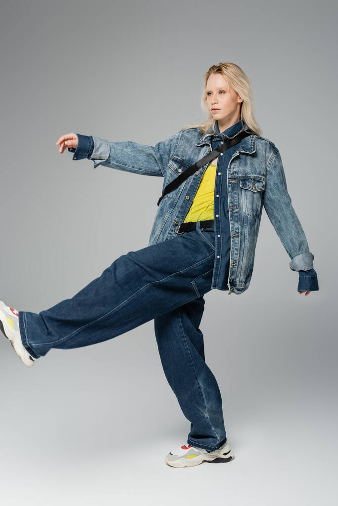 full length of young blonde woman in trendy denim outfit posing while walking on grey  - Foto, afbeelding