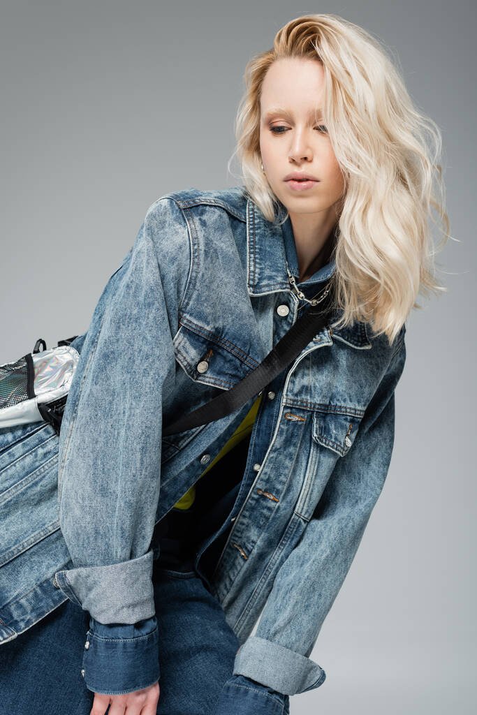 молодая блондинка в стильной джинсовой куртке и ременной сумке позирует изолированно на сером  - Фото, изображение