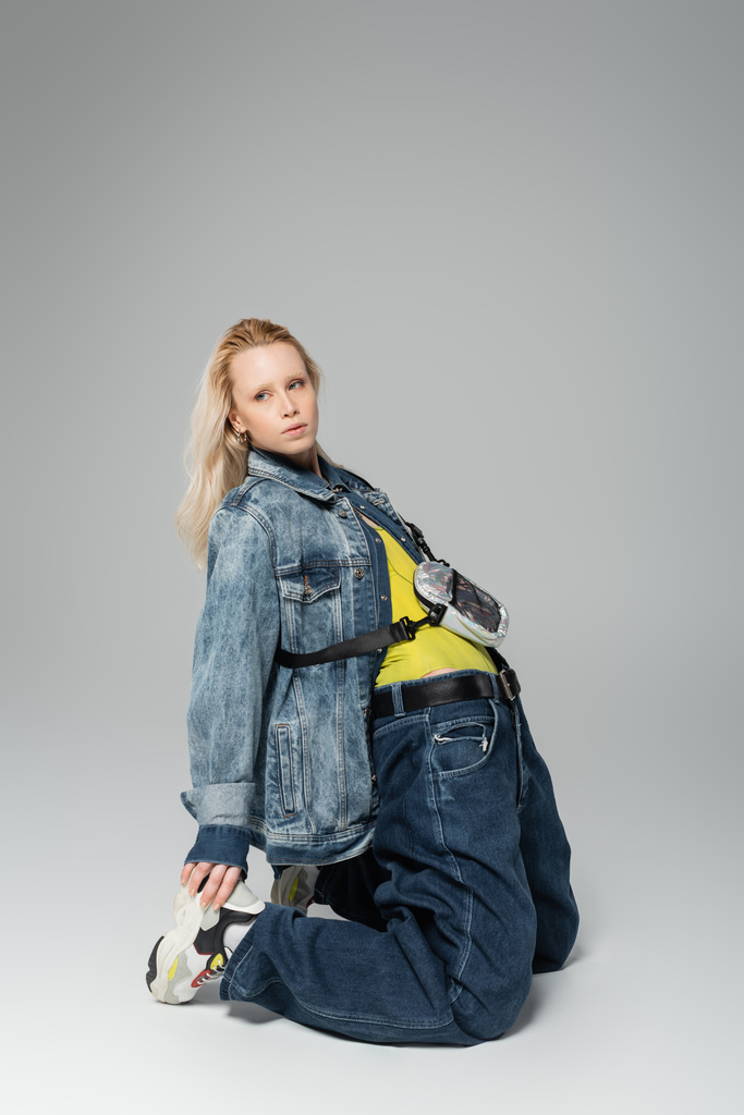 full length of young blonde woman in blue denim outfit and trendy sneakers posing on grey  - 写真・画像
