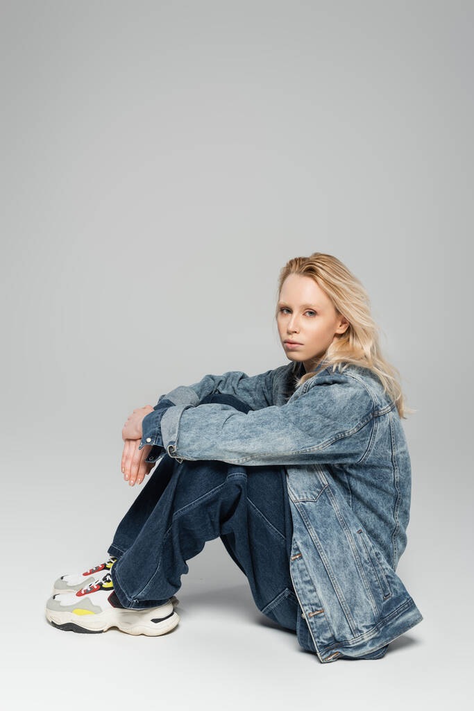 full length of blonde albino woman in total denim outfit and trendy sneakers sitting on grey  - Photo, image