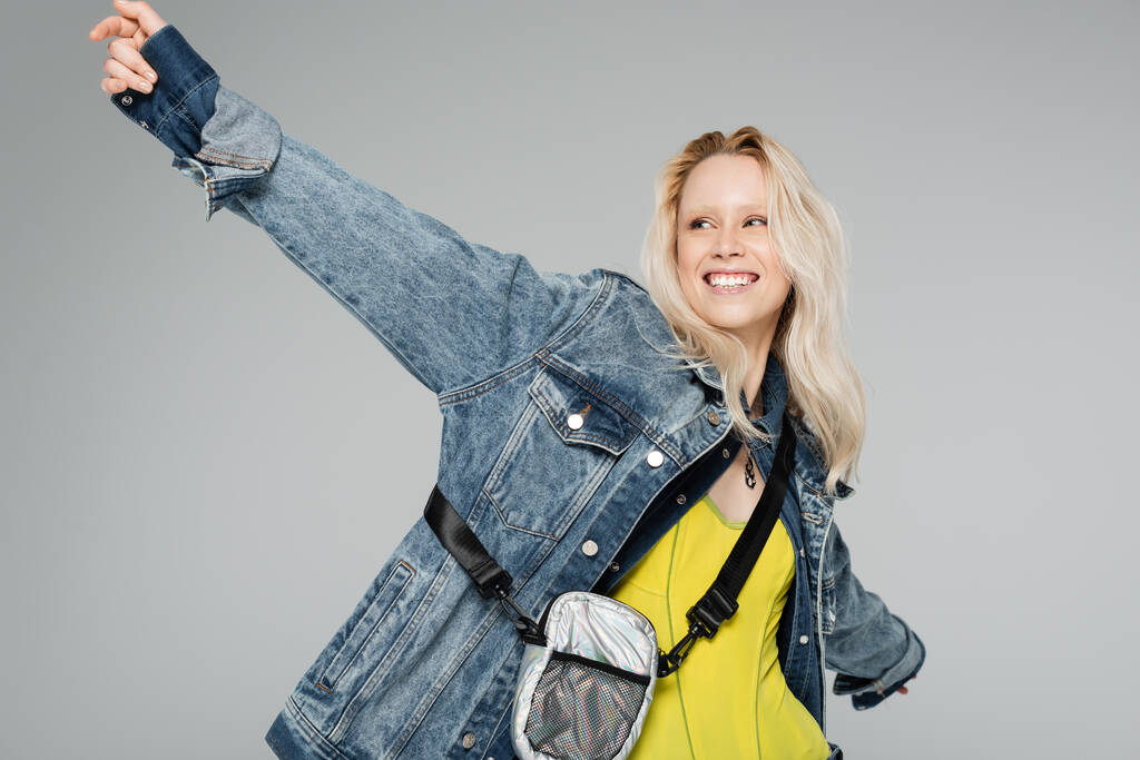 happy blonde woman in stylish denim jacket with belt bag standing with outstretched hands isolated on grey  - Photo, Image