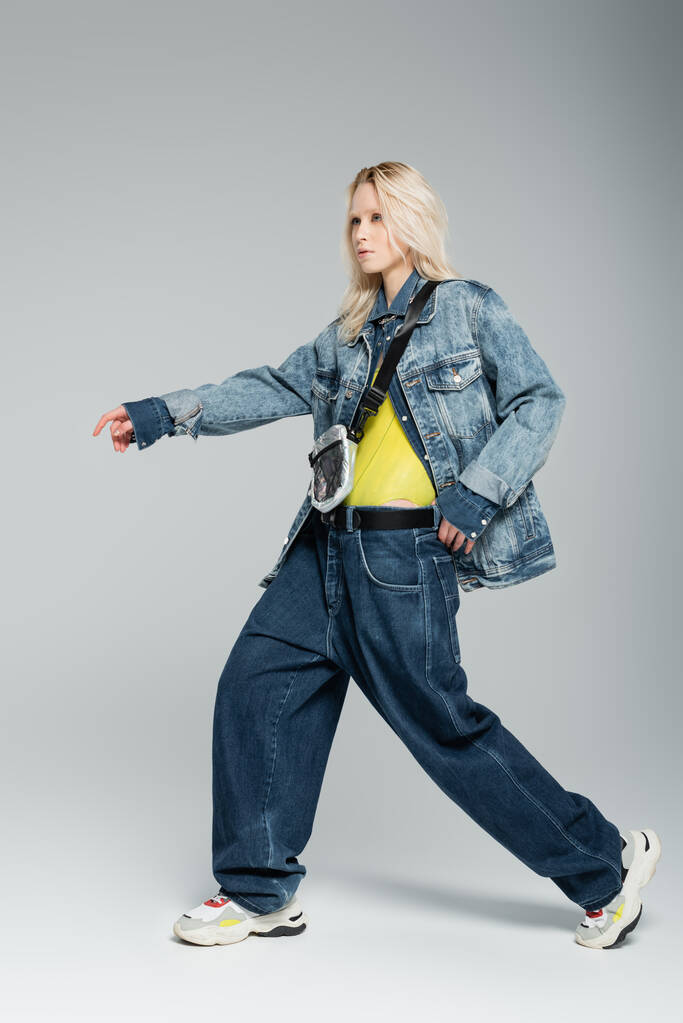 full length of young blonde woman in blue denim outfit and trendy sneakers posing with outstretched hand on grey  - Fotografie, Obrázek