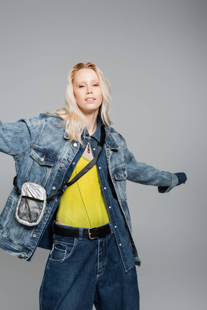 young blonde woman in denim blue jacket and belt bag posing with outstretched hand isolated on grey  - Fotó, kép