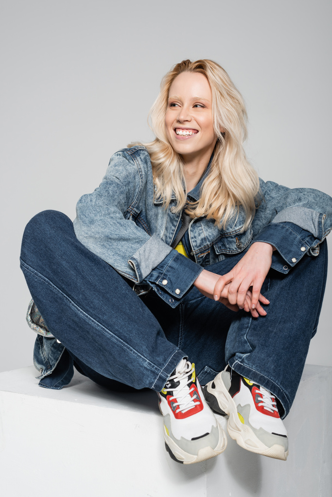 полная длина счастливая блондинка в синем джинсовом наряде и модные кроссовки, сидящие на белом кубе изолированы на сером  - Фото, изображение