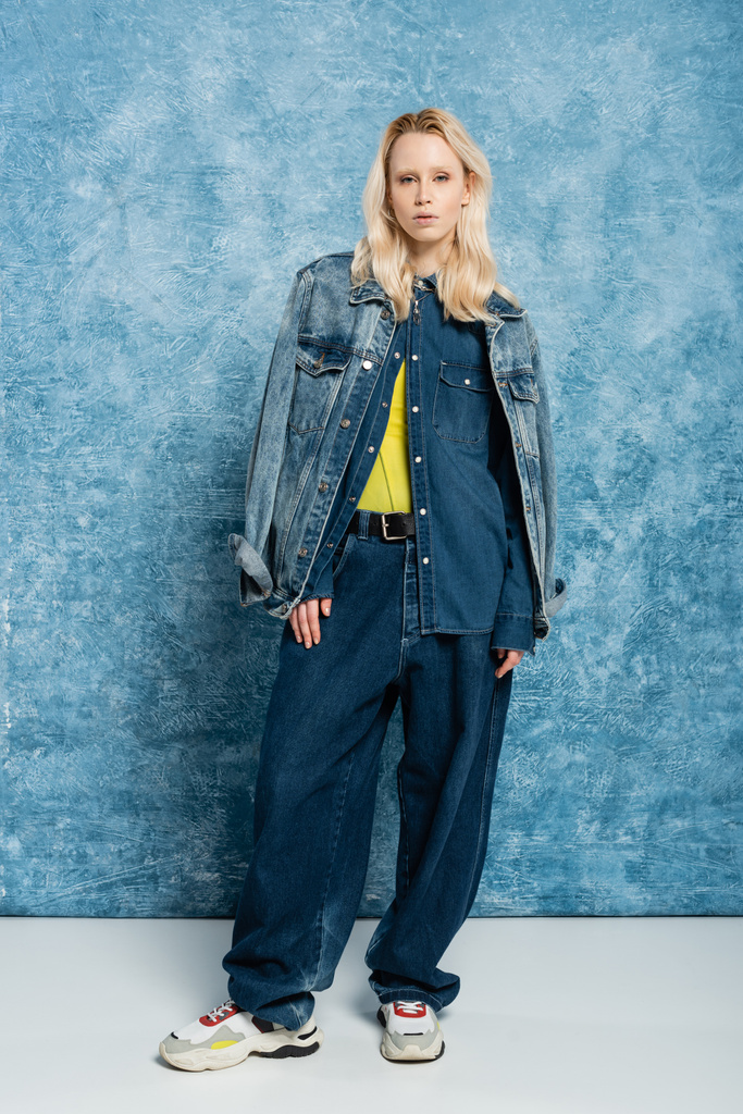 full length of blonde woman in denim outfit posing near blue textured background   - Foto, Bild