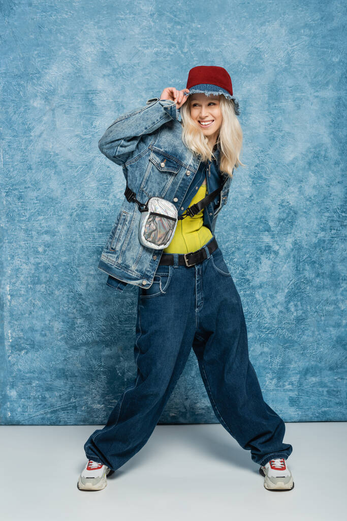 full length of happy blonde woman in denim outfit adjusting panama hat near blue textured background   - 写真・画像