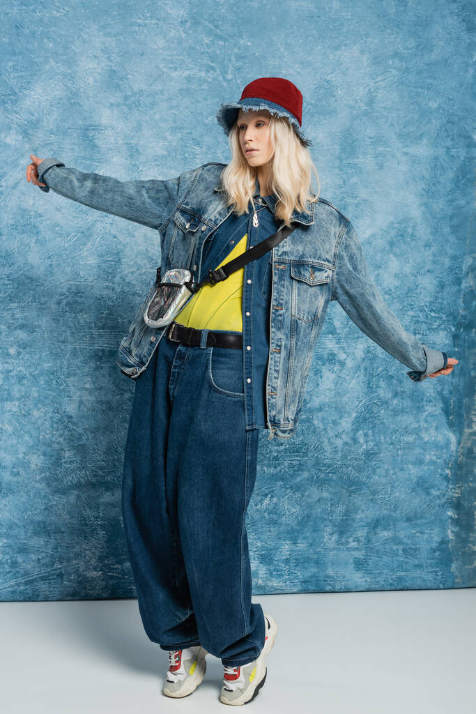 full length of blonde woman in denim jacket and panama hat posing with outstretched hands near blue textured background   - 写真・画像