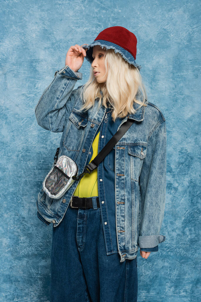 блондинка в джинсовой куртке регулирует модную панамскую шляпу возле синего текстурированного фона   - Фото, изображение