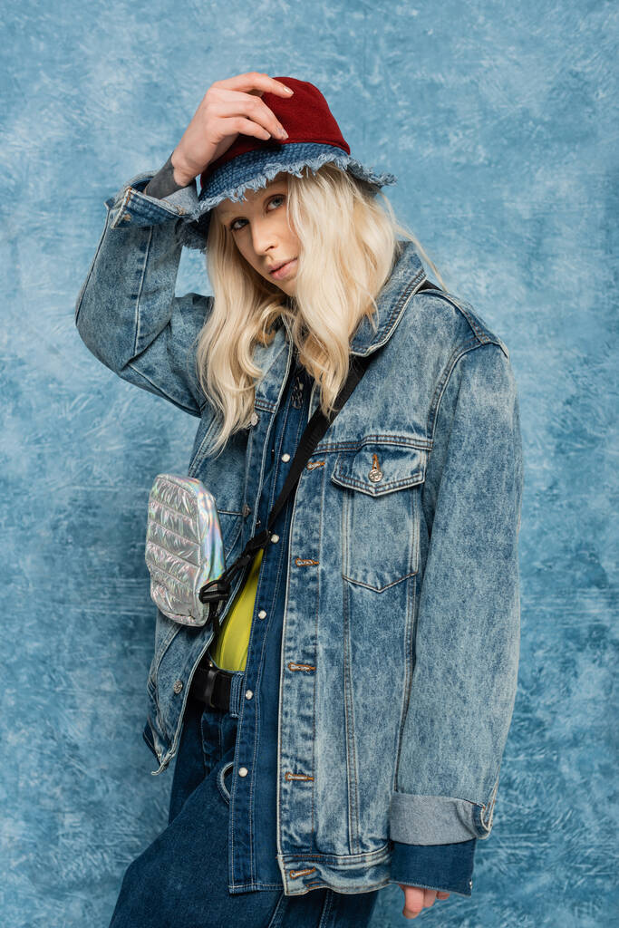 blondi nainen denim takki ja panama hattu katselee kameraa lähellä sininen kuvioitu tausta   - Valokuva, kuva