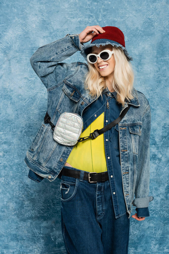 весела блондинка в джинсовому вбранні позує в панамському капелюсі і сонцезахисних окулярах біля синього текстурованого фону
   - Фото, зображення