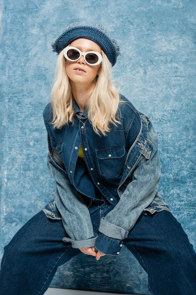 young blonde woman in denim outfit posing in panama hat and sunglasses near blue textured background   - 写真・画像