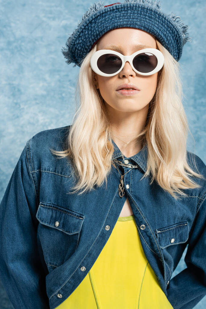 young blonde woman in denim panama hat and sunglasses posing near blue textured background   - Fotó, kép