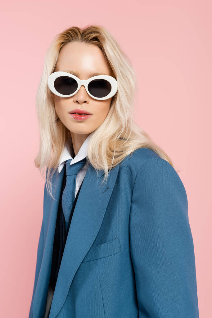 young blonde woman in blue blazer and sunglasses looking at camera isolated on pink - Foto, Imagen