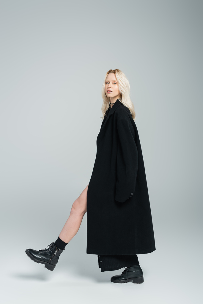 longitud completa de la joven rubia en abrigo negro caminando y mirando a la cámara en gris  - Foto, Imagen