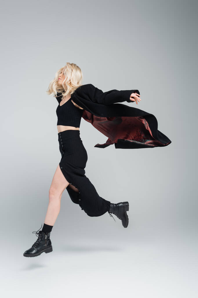 full length of blonde and young woman in stylish black outfit jumping on grey  - Fotografie, Obrázek