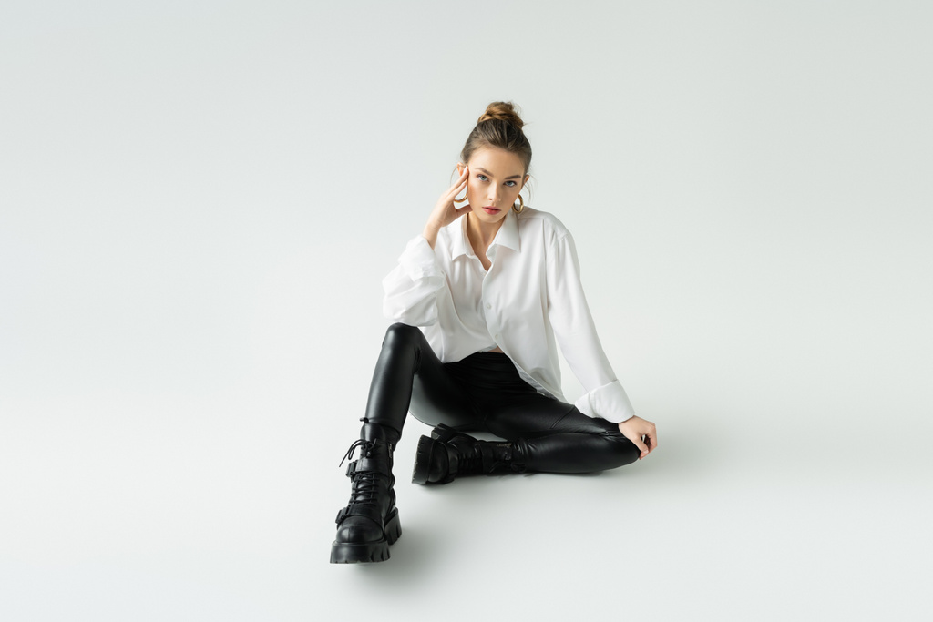 longitud completa de modelo joven en pantalones de látex negro y botas ásperas sentado sobre fondo gris - Foto, imagen