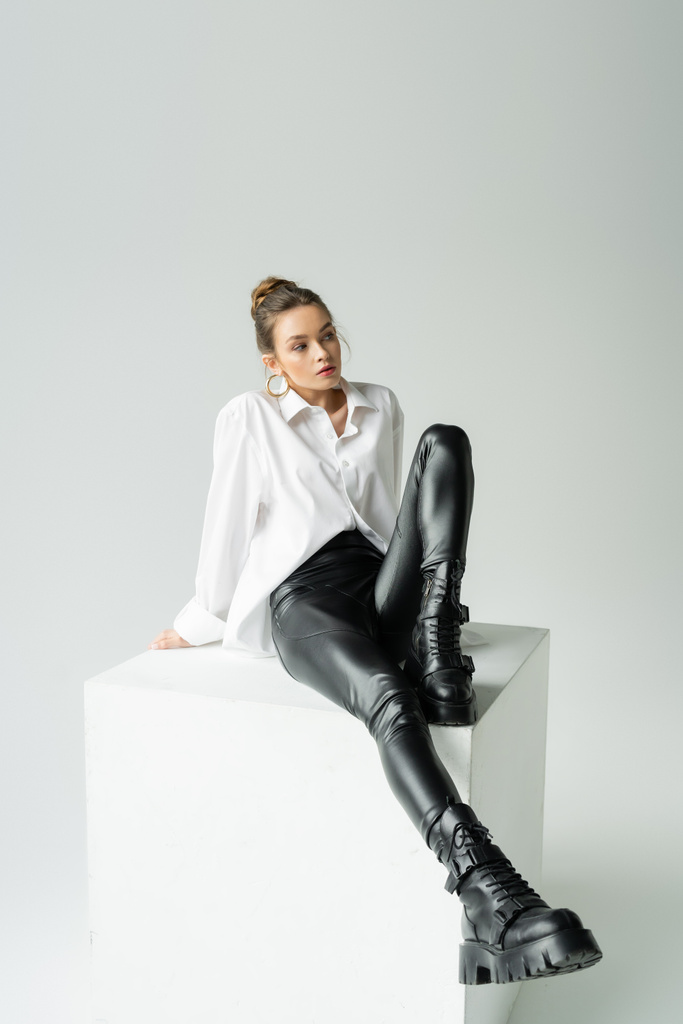 full length of woman in black latex pants and rough boots sitting on white cube on grey background - Valokuva, kuva