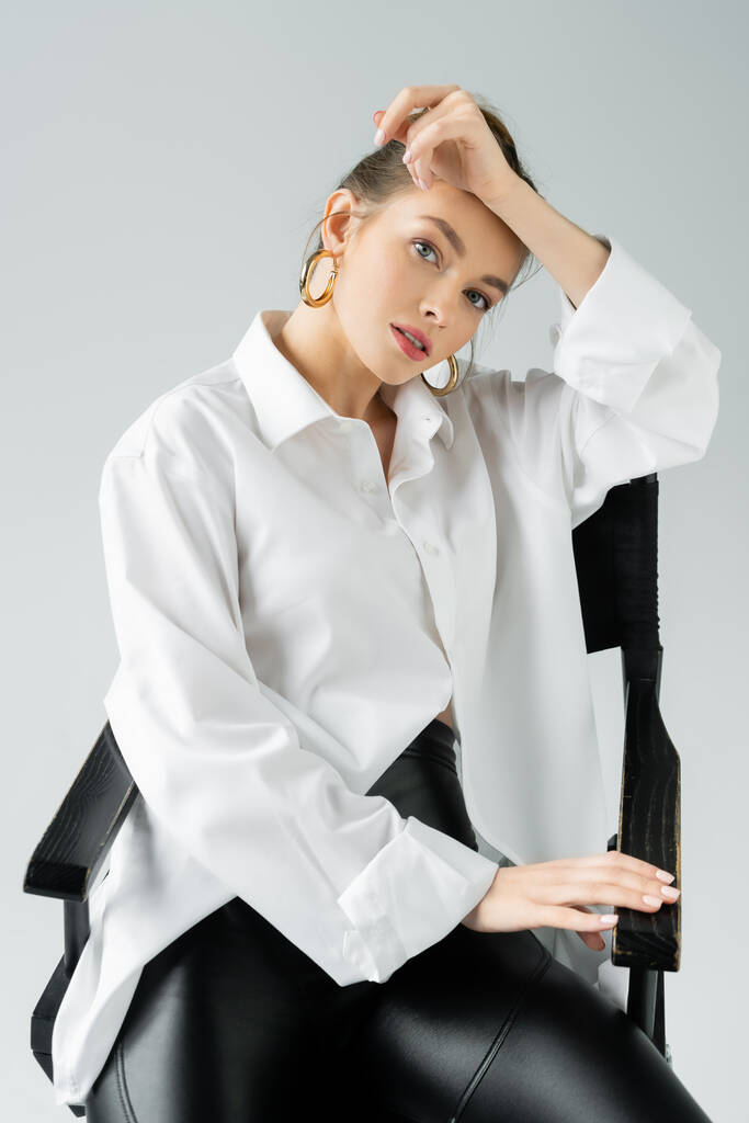 stylish woman in hoop earrings and white oversize shirt posing on chair and looking at camera isolated on grey - Photo, Image