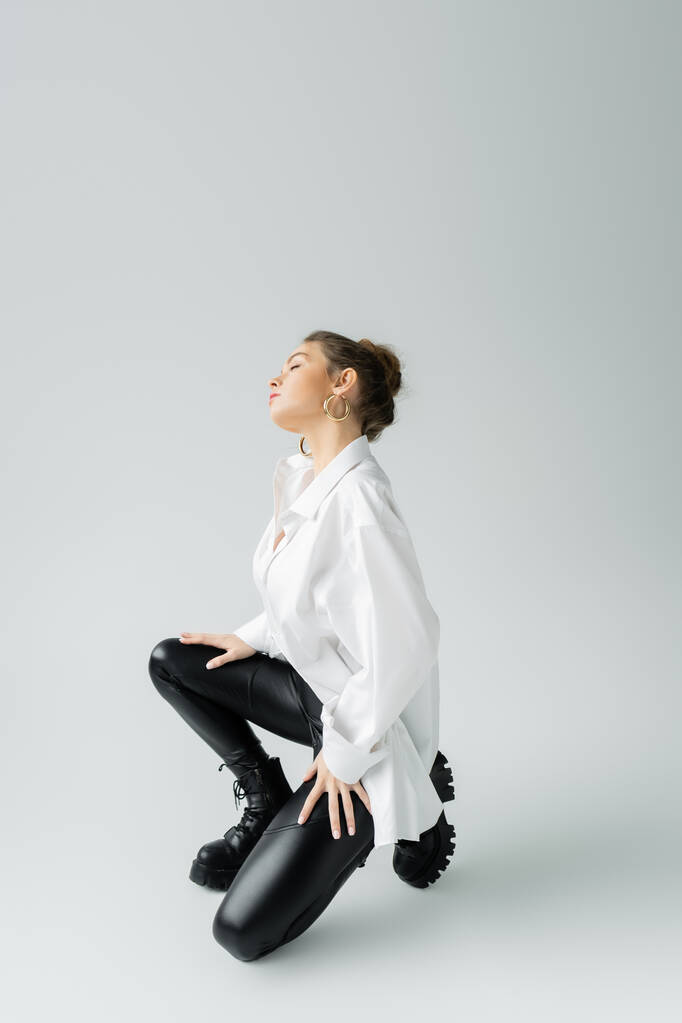 full length of stylish woman in white oversize shirt and black tight pants posing on grey background - Foto, immagini